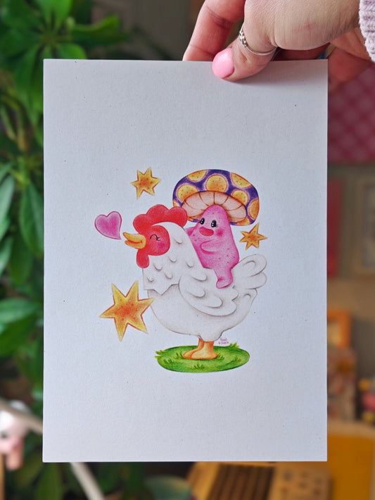 A hand holds a white art print featuring a cute colour pencil illustration of a white chicken with a pink mushroom with a purple and orange mushroom cap, yellow stars, and a green patch of grass, against a blurred indoor background with plants and objects.